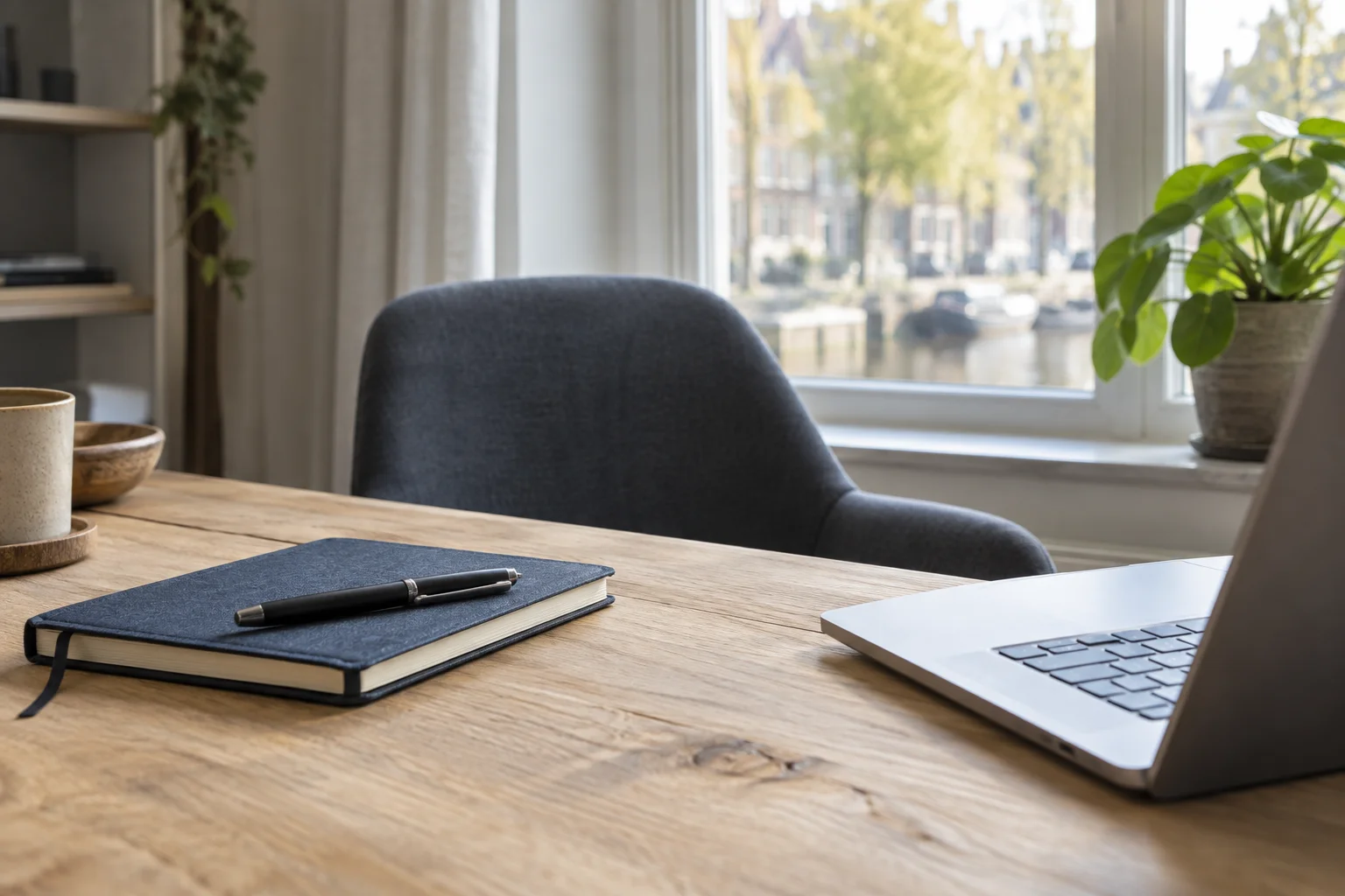 Een rustige tafel bij het raam met een notitieboek, laptop en lege stoel.